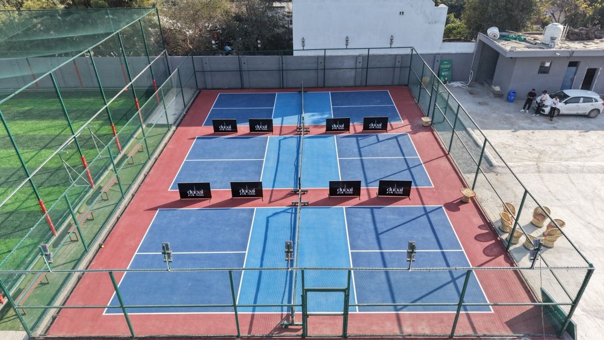Aerial view of the Dubai Fieldz pickleball courts: blue hardcourts with white lines and Dubai Fieldz banners along the net, the green turf cage to the left