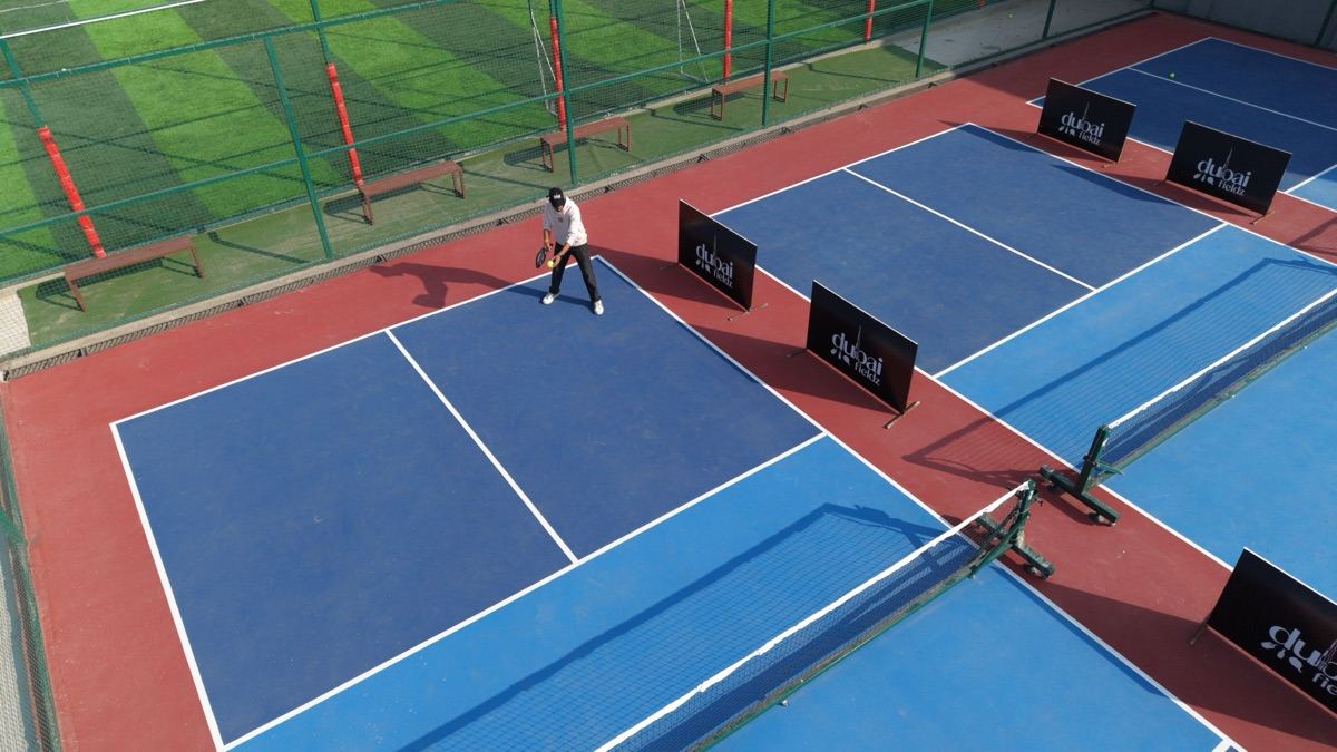 A player serves on a blue pickleball hardcourt at Dubai Fieldz, Dubai Fieldz banners between the courts