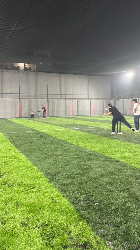 Box cricket at night under floodlights: a batter plays a shot at the stumps on striped turf, the netted cage behind
