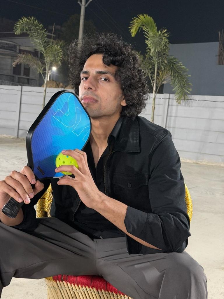 A player in black holds a blue pickleball paddle and a yellow ball, palms and a white wall behind him at dusk