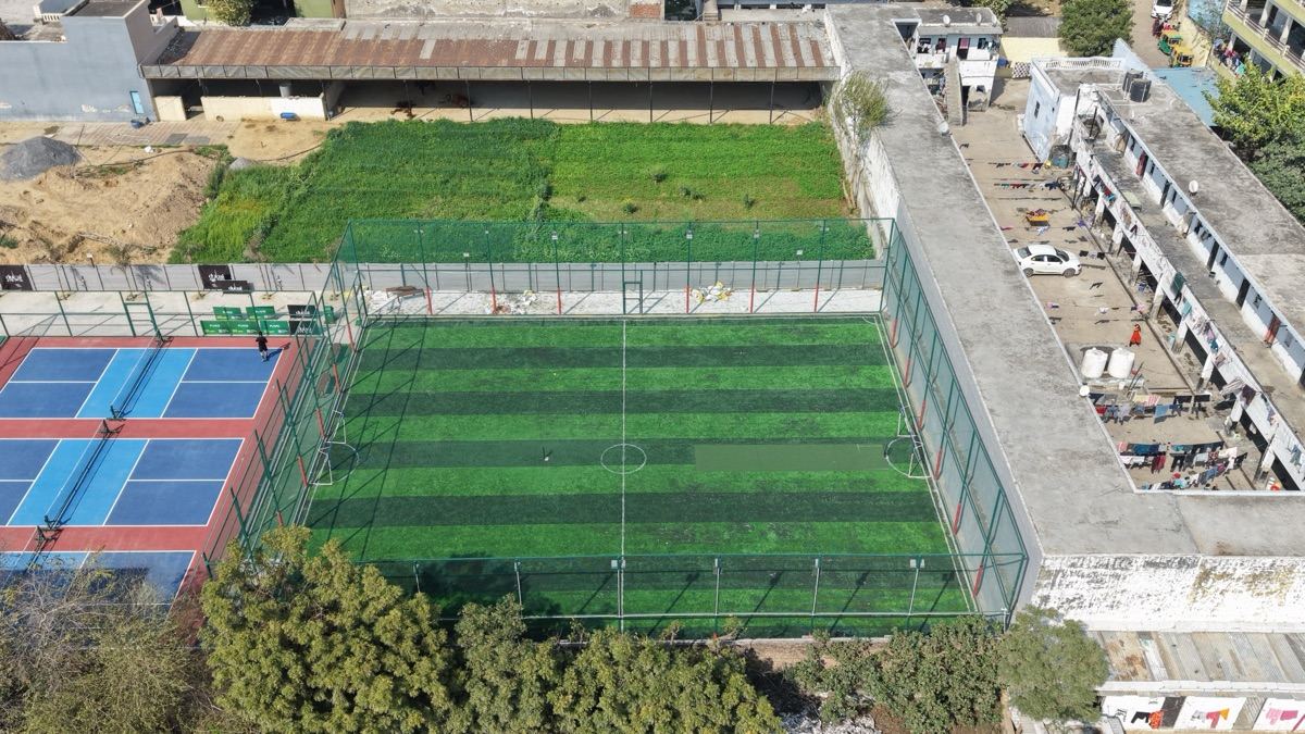 Aerial view of the Dubai Fieldz football turf: striped artificial grass inside a tall green cage, with the pickleball courts beside it